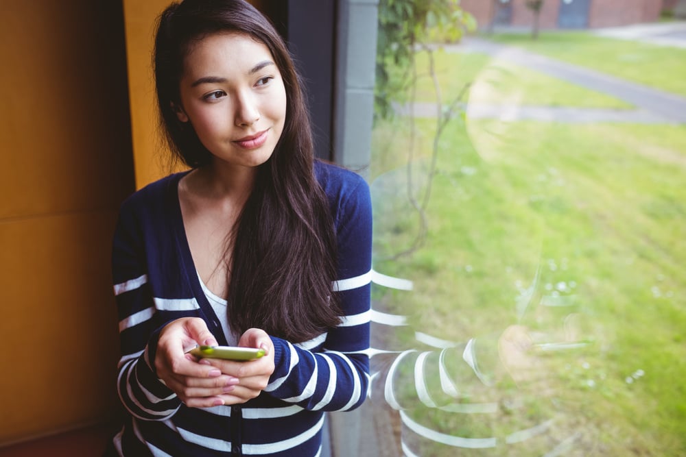 Smiling student with smartphone and looking through the window at the university-1